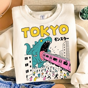 Può includere: Felpa color sabbia con una grafica di un mostro turchese che mangia un treno rosa a Tokyo. La parola "TOKYO" è in lettere gialle a blocchi sopra il mostro. È presente anche la parola giapponese per "mostro".