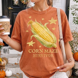 May include: A rust-colored short-sleeve t-shirt featuring a graphic of a yellow corn cob surrounded by yellow stars. The text "CORN MAZE SURVIVOR" is printed in yellow below the corn. The shirt has a crew neckline and a relaxed fit.
