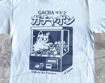 May include: A light blue t-shirt featuring a dark blue graphic of a cat inside a gacha machine. The text reads GACHA マシン ガチャポン and Unbox the Flavors of Japan. The design includes a cat reaching for spherical capsules inside the machine.