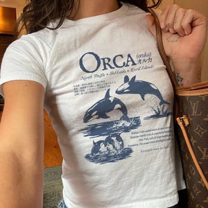 Puede incluir: Una camiseta blanca de manga corta con un estampado gráfico en azul oscuro de orcas. El diseño incluye el texto ORCA, caracteres japoneses y detalles descriptivos sobre el hábitat, comportamiento, dieta y reputación del mamífero marino.