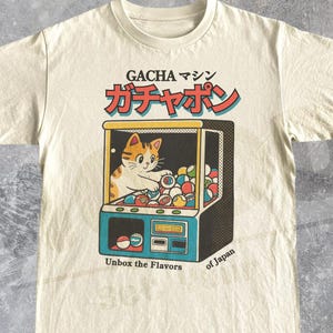 Puede incluir: Camiseta color crema con un gráfico retro de un gato en una máquina gacha. La máquina es azul, amarilla y negra, con texto japonés arriba y la frase "Unbox the Flavors of Japan" abajo.