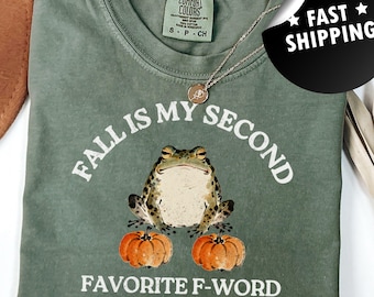 May include: Sage green t-shirt with the text "FALL IS MY SECOND FAVORITE F-WORD THE FIRST ONE IS FROG". The graphic features a frog sitting on two orange pumpkins. A silver necklace is draped across the shirt.
