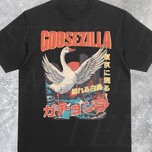 Puede incluir: Camiseta negra con un gráfico de un ganso con las alas extendidas, etiquetado como "GOOSEZILLA" en naranja. El diseño también incluye texto japonés y un paisaje urbano. El nombre de la tienda Etsy "SetYourMoodTee" está en la parte inferior.