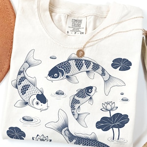 Puede incluir: Camiseta color crema con una ilustración azul marino de peces koi nadando entre nenúfares y piedras. Un collar dorado con un colgante descansa sobre la camiseta. La camiseta tiene una etiqueta Comfort Colors.