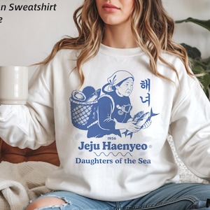 Op de afbeelding: Wit Gildan sweatshirt met een blauwe afbeelding van een vrouw met een mand en vis, met Koreaanse en Engelse tekst: "Jeju Haenyeo, Daughters of the Sea." De sweatshirt heeft een ronde hals.