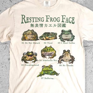 May include: An ivory short-sleeve t-shirt featuring the text Resting Frog Face and Japanese characters above seven illustrated frogs. Each frog is numbered and labeled with phrases including Do Not Disturb, Tired, I Need Coffee, Absolutely Not, Overstimulated, No Thanks, and Buffering.