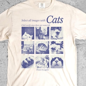 May include: Ivory-colored t-shirt with a blue graphic of cat images and the text "Select all images with Cats." The shirt also includes the text "Click verify once there are none left" and "Please try again."