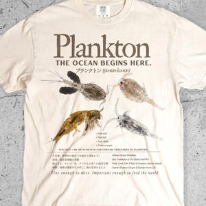 May include: An ivory short-sleeve t-shirt featuring a graphic print of four plankton organisms. The text reads Plankton, The Ocean Begins Here, and includes scientific facts in English and Japanese, plus the phrase Tiny enough to miss.