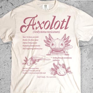 Könnte beinhalten: Ein elfenbeinfarbenes T-Shirt mit einem rosa Grafikdruck von Axolotln und dem Text Axolotl Ambystoma mexicanum, State: No brain, just smile, Routine: Eat, float, repeat, Defense: Being too cute, Reputation: Internet favorite, Skill: Regrowing problems away.