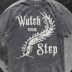 May include: A charcoal grey short-sleeve t-shirt featuring a white graphic of a centipede curving through the text "Watch YOUR Step" printed in a gothic-style font. The shirt has a crew neckline and a faded, washed-out fabric appearance.