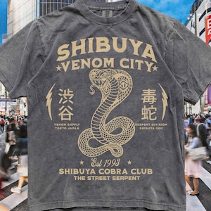 May include: A charcoal grey short-sleeve t-shirt featuring a beige graphic of a cobra snake. The text reads SHIBUYA VENOM CITY, SHIBUYA COBRA CLUB, THE STREET SERPENT, EST 1993, VENOM SUPPLY TOKYO JAPAN, SERPENT DIVISION SHIBUYA 1993, and Japanese characters.
