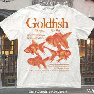 Peut inclure: T-shirt blanc aux couleurs confort avec un motif orange de poissons rouges et le mot "Goldfish" en orange. Le texte comprend également des caractères japonais et du texte supplémentaire sur les poissons.