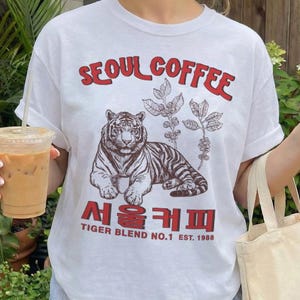 Peut inclure: Un t-shirt blanc avec un motif représentant un tigre et des branches de caféier. Le texte indique SEOUL COFFEE en rouge, accompagné de caractères coréens et de la mention TIGER BLEND NO.1 EST. 1988. Il est porté avec un short rayé bleu clair.