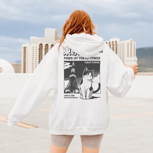 May include: White hoodie with a graphic on the back featuring a black and white cat, the Tokyo Tower, and the text "PAWS AT TOKYO TOWER" and "TOKYO TOWER SINCE 1958". The hoodie has a drawstring hood and long sleeves.