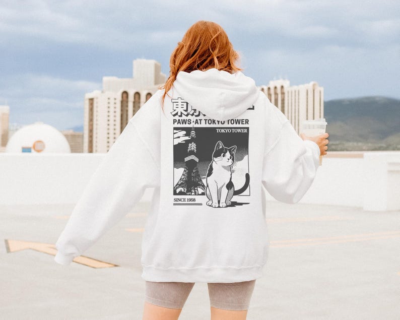 May include: White hoodie with a graphic on the back featuring a black and white cat, the Tokyo Tower, and the text "PAWS AT TOKYO TOWER" and "TOKYO TOWER SINCE 1958". The hoodie has a drawstring hood and long sleeves.