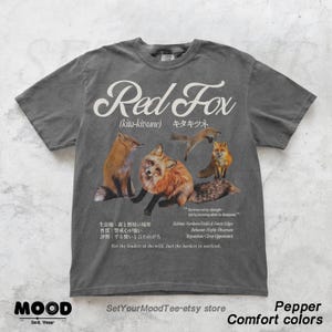 K&ouml;nnte beinhalten: Graues T-Shirt mit einem Aufdruck von Rotf&uuml;chsen und dem Text "Red Fox" in Englisch und Japanisch. Der Entwurf enth&auml;lt den Satz "Not the loudest in the wild. Just the hardest to outthink." Teil der "Comfort colors"-Kollektion.