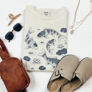 May include: Cream-colored sweatshirt with a koi fish design, sunglasses, a brown sling bag, gold hair clip, gold necklaces, and beige slip-on sandals. The koi fish are in shades of gray and blue.