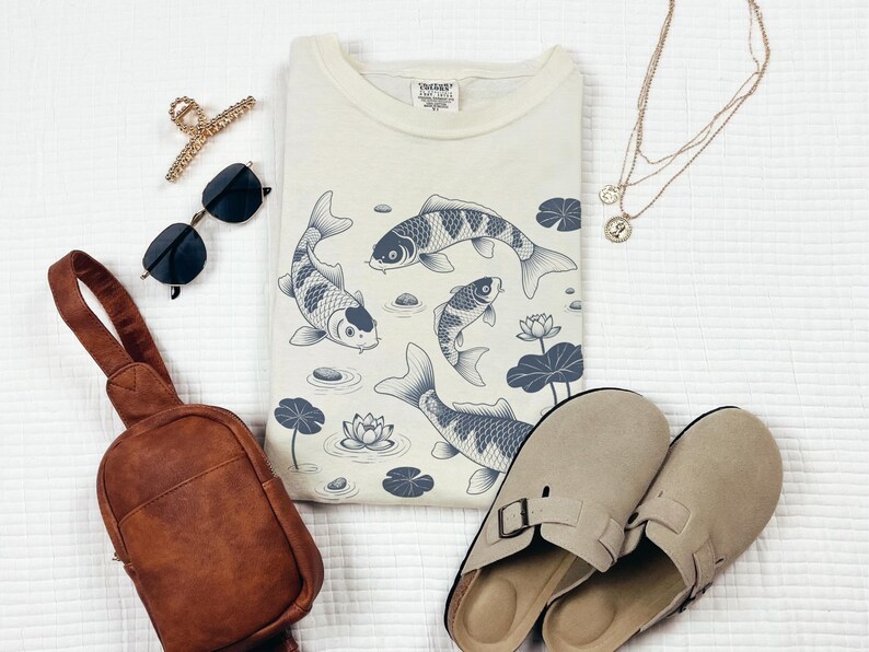 May include: Cream-colored sweatshirt with a koi fish design, sunglasses, a brown sling bag, gold hair clip, gold necklaces, and beige slip-on sandals. The koi fish are in shades of gray and blue.