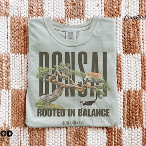 May include: A light green t-shirt with a bonsai tree graphic and the word "BONSAI" in large, dark letters. The shirt also features the text "ROOTED IN BALANCE" and Japanese text. Two cats are in the tree.