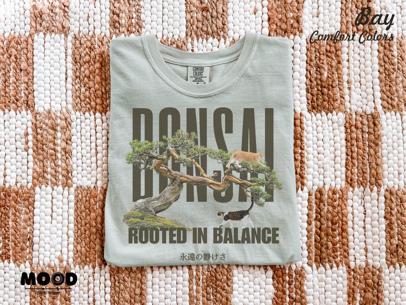 May include: A light green t-shirt with a bonsai tree graphic and the word "BONSAI" in large, dark letters. The shirt also features the text "ROOTED IN BALANCE" and Japanese text. Two cats are in the tree.
