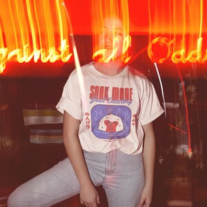 May include: Cream-colored t-shirt with the text "SOAK MODE just existing" and a graphic of a monkey in a hot spring. The shirt is paired with light blue jeans. The background features blurred neon lights.