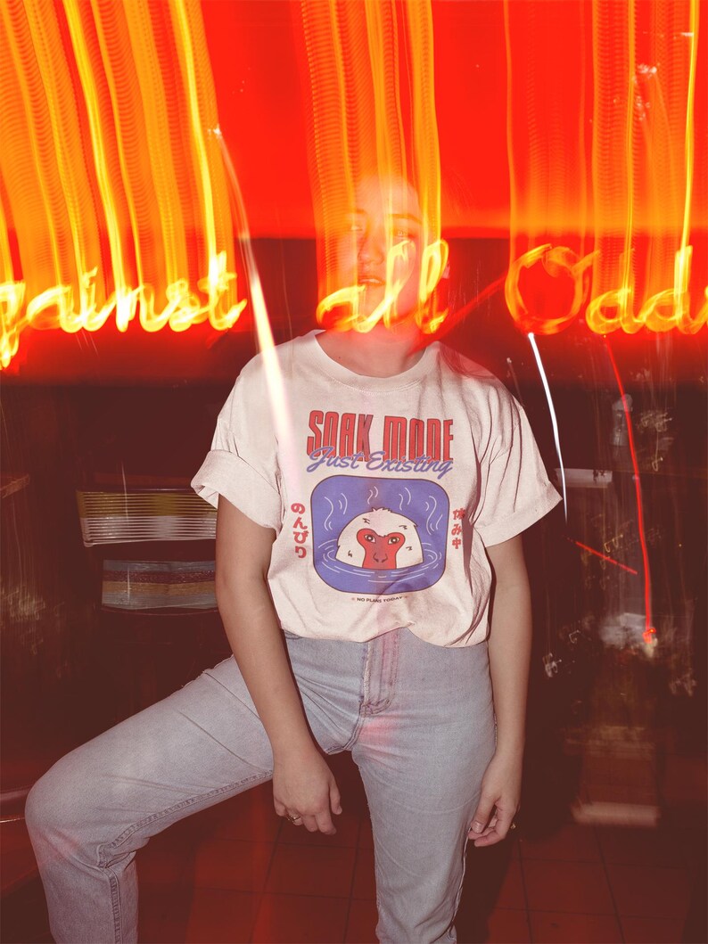 May include: Cream-colored t-shirt with the text "SOAK MODE just existing" and a graphic of a monkey in a hot spring. The shirt is paired with light blue jeans. The background features blurred neon lights.