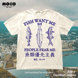 May include: Ivory-colored t-shirt with the text "FISH WANT ME PEOPLE FEAR ME" and three blue fish illustrations. Below the text are Japanese characters. The shirt also has the text "Comfort colors" and the Etsy store name.