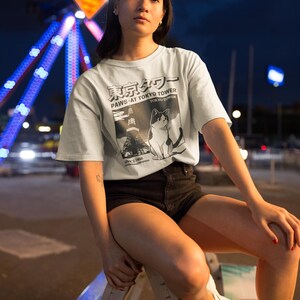 以下が含まれることがあります： 猫のグラフィックと「PAWS AT TOKYO TOWER」の文字が入った水色のTシャツ。シャツは黒いショートパンツと組み合わせています。背景には夜の観覧車があります。