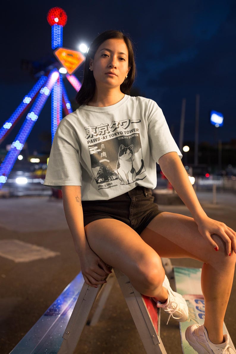 以下が含まれることがあります： 猫のグラフィックと「PAWS AT TOKYO TOWER」の文字が入った水色のTシャツ。シャツは黒いショートパンツと組み合わせています。背景には夜の観覧車があります。