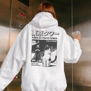 May include: White hoodie with a graphic on the back depicting the Tokyo Tower and a black and white cat. The text reads "Paws at Tokyo Tower" and "Since 1958". The hoodie has a drawstring hood and long sleeves.
