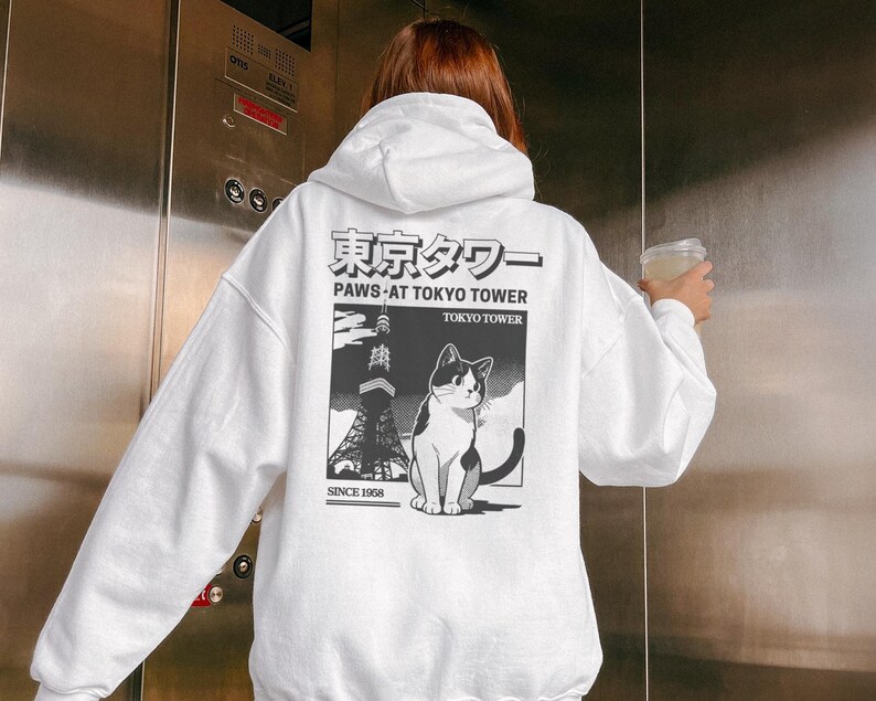 May include: White hoodie with a graphic on the back depicting the Tokyo Tower and a black and white cat. The text reads "Paws at Tokyo Tower" and "Since 1958". The hoodie has a drawstring hood and long sleeves.