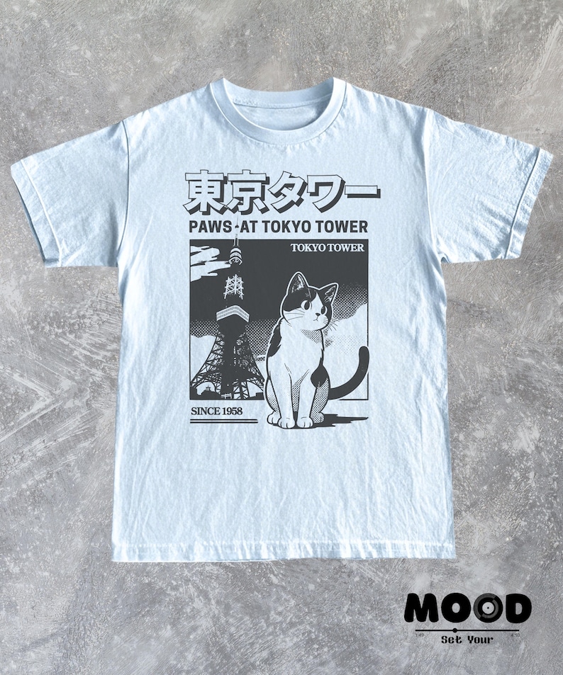 以下が含まれることがあります： 東京タワーの前にいる猫の白黒グラフィックが特徴のライトブルーTシャツ。「Paws at Tokyo Tower」と「Since 1958」の文字入り。クルーネックと半袖のデザインです。