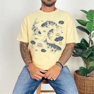 May include: A light yellow t-shirt featuring a koi fish design. The graphic shows several koi fish swimming among lily pads and lotus flowers. The shirt is a relaxed fit and has short sleeves.