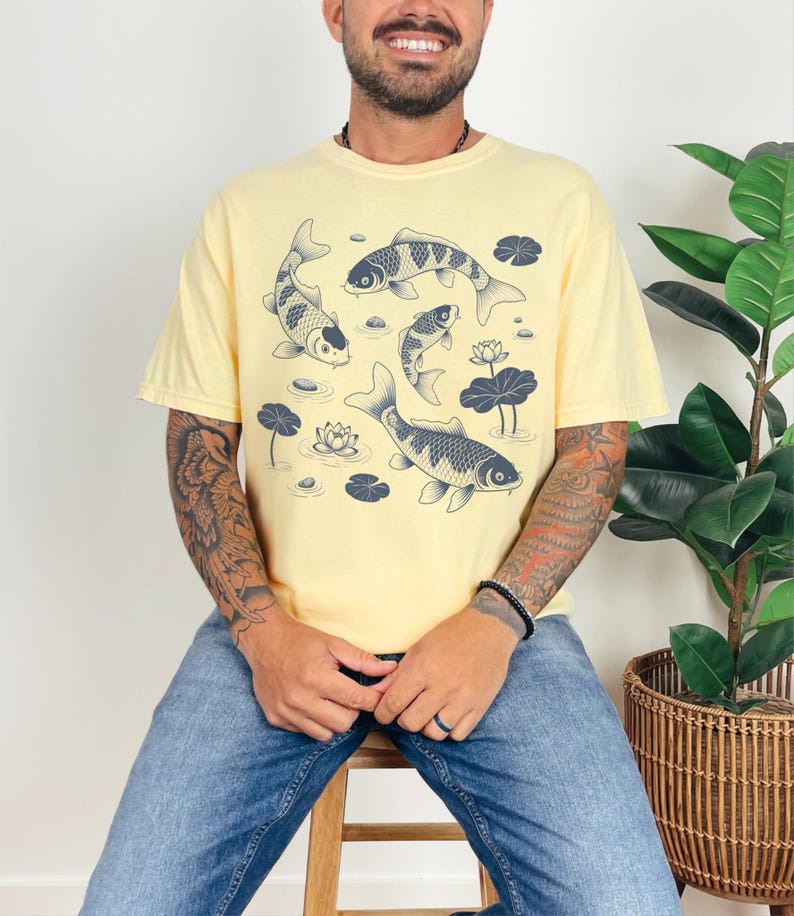 May include: A light yellow t-shirt featuring a koi fish design. The graphic shows several koi fish swimming among lily pads and lotus flowers. The shirt is a relaxed fit and has short sleeves.