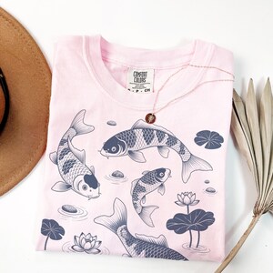May include: A light pink t-shirt with a crew neck featuring a koi fish design in shades of gray. The shirt has a Comfort Colors tag and is paired with a gold necklace. The design includes water lilies and stones.