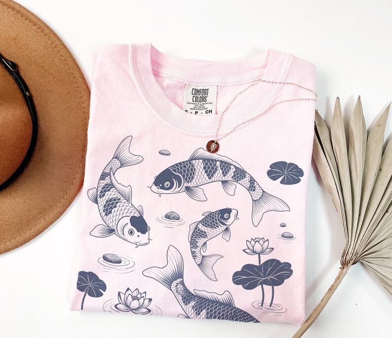 May include: A light pink t-shirt with a crew neck featuring a koi fish design in shades of gray. The shirt has a Comfort Colors tag and is paired with a gold necklace. The design includes water lilies and stones.