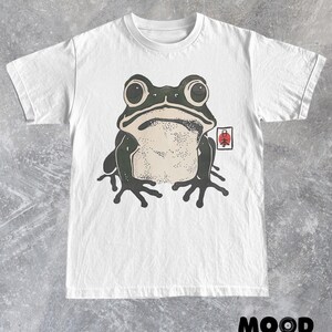 May include: White t-shirt featuring a large, stylized frog illustration in black and beige. The frog has large eyes and is centered on the shirt. A red and white Japanese symbol is on the right. The text "MOOD Set Your" is at the bottom.