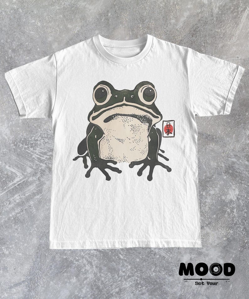 May include: White t-shirt featuring a large, stylized frog illustration in black and beige. The frog has large eyes and is centered on the shirt. A red and white Japanese symbol is on the right. The text "MOOD Set Your" is at the bottom.