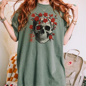 May include: Sage green t-shirt featuring a detailed skull graphic adorned with red cherry blossoms. The skull has one red eye and is framed by the flowers. Japanese text is printed below the skull. The shirt has short sleeves.