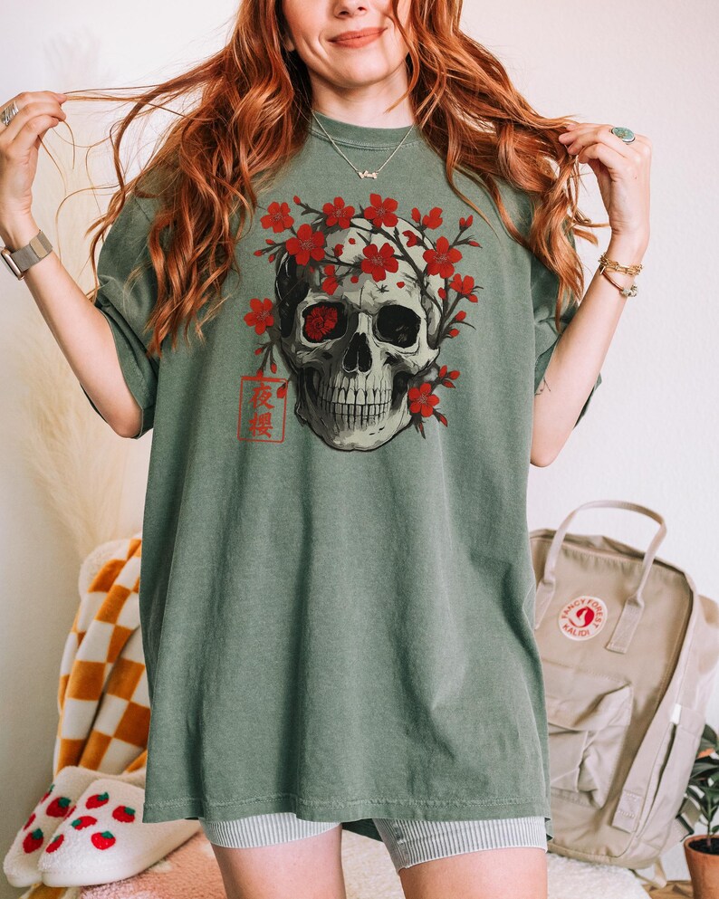 May include: Sage green t-shirt featuring a detailed skull graphic adorned with red cherry blossoms. The skull has one red eye and is framed by the flowers. Japanese text is printed below the skull. The shirt has short sleeves.