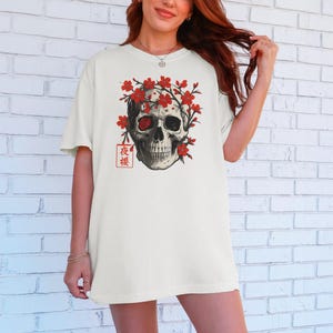 May include: Off-white t-shirt featuring a detailed skull graphic adorned with red flowers. The design includes Japanese text in a red square. The skull has a red eye and is surrounded by red blossoms, creating a striking contrast.