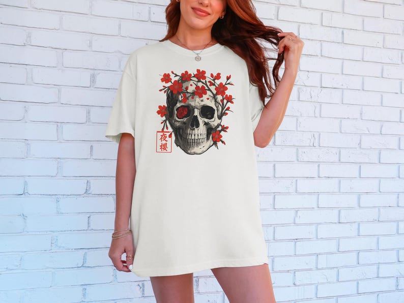 May include: Off-white t-shirt featuring a detailed skull graphic adorned with red flowers. The design includes Japanese text in a red square. The skull has a red eye and is surrounded by red blossoms, creating a striking contrast.