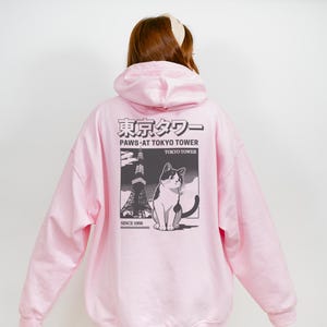 May include: Pale pink hoodie with a graphic on the back depicting a cat, the Tokyo Tower, and Japanese text. The graphic is in monochrome. The hoodie has a hood and long sleeves.