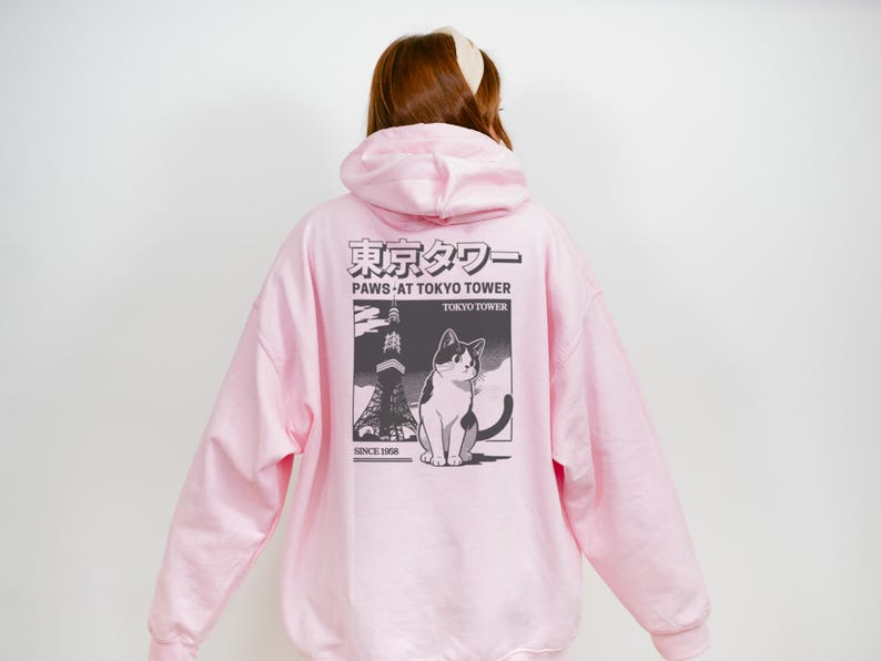 May include: Pale pink hoodie with a graphic on the back depicting a cat, the Tokyo Tower, and Japanese text. The graphic is in monochrome. The hoodie has a hood and long sleeves.