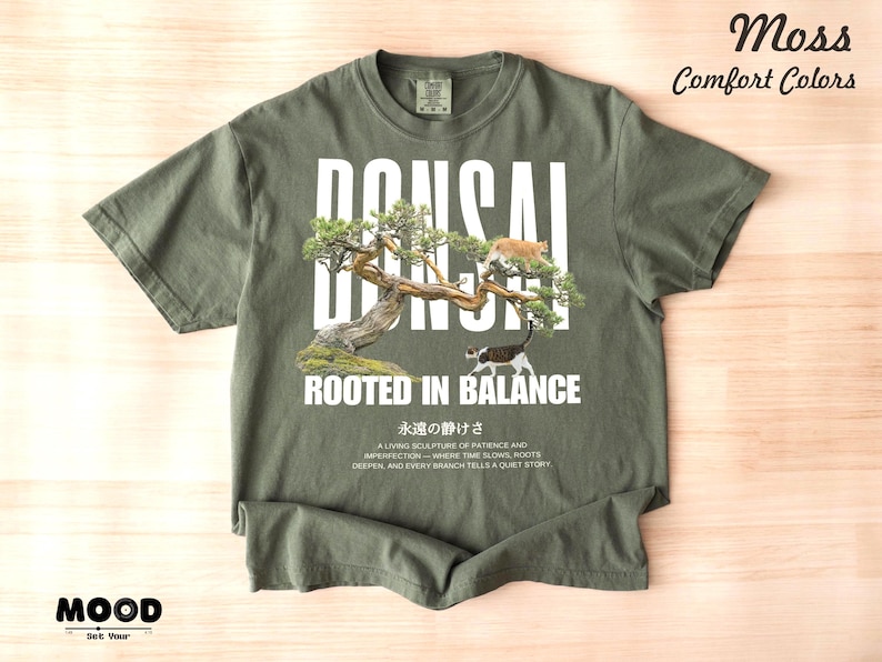 May include: Moss green t-shirt with a bonsai tree graphic and the text "BONSAI ROOTED IN BALANCE". The design includes two cats. Additional text at the bottom reads "A living sculpture of patience..."