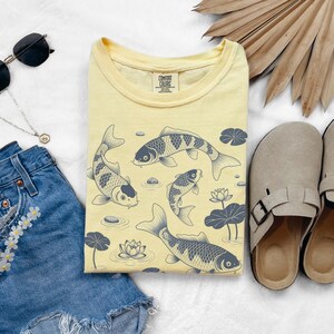 May include: A light yellow t-shirt featuring a koi fish design with water lilies. The shirt is paired with denim shorts, sunglasses, a necklace, and beige sandals. The shirt has the text "COMFORT COLORS".