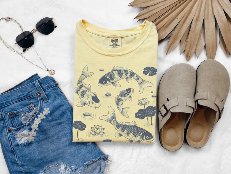 May include: A light yellow t-shirt featuring a koi fish design with water lilies. The shirt is paired with denim shorts, sunglasses, a necklace, and beige sandals. The shirt has the text "COMFORT COLORS".