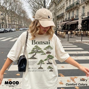 May include: Ivory-colored t-shirt with a Bonsai tree graphic and text. The shirt features a crew neck and short sleeves. A beige baseball cap with a black logo is worn. A black purse with a chain strap is also visible.