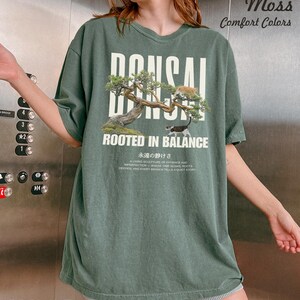 May include: A moss green t-shirt with the word "BONSAI" in white block letters above an illustration of a bonsai tree and two cats. The text "ROOTED IN BALANCE" is below the illustration. The shirt also has Japanese text.