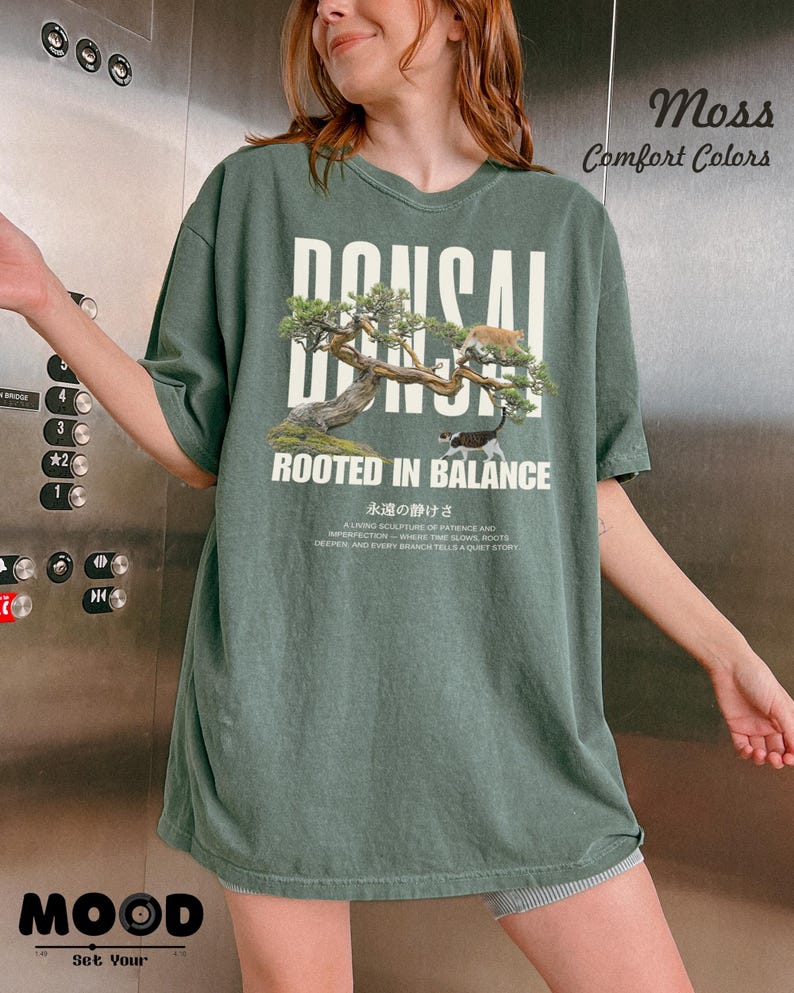 May include: A moss green t-shirt with the word "BONSAI" in white block letters above an illustration of a bonsai tree and two cats. The text "ROOTED IN BALANCE" is below the illustration. The shirt also has Japanese text.